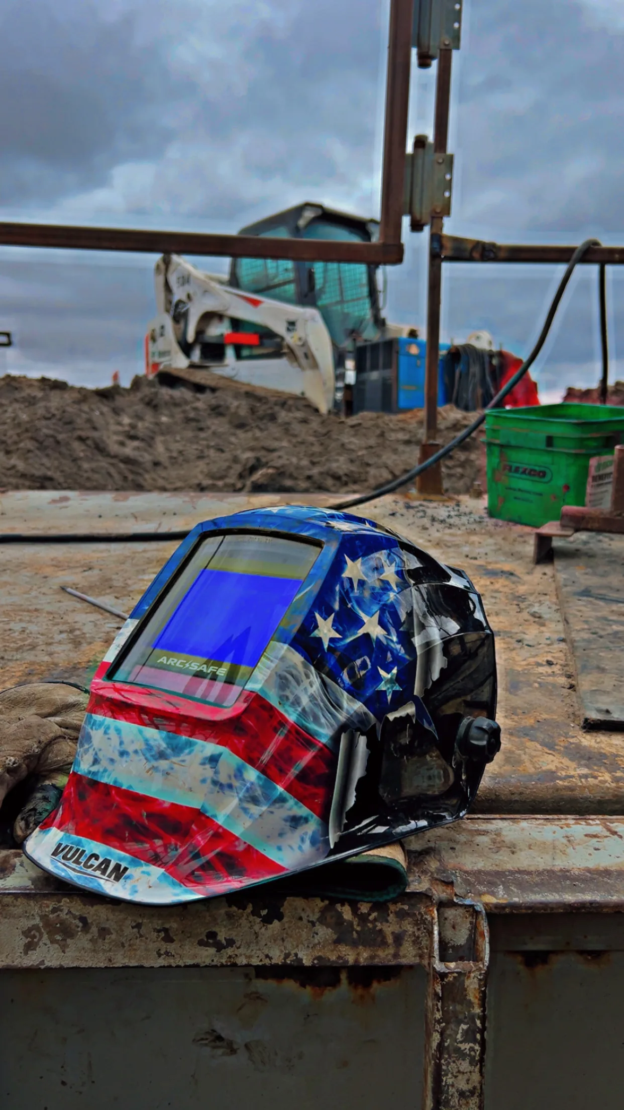 American flag welding helmet on a rusty steel beam with Bobcat skid steer in background