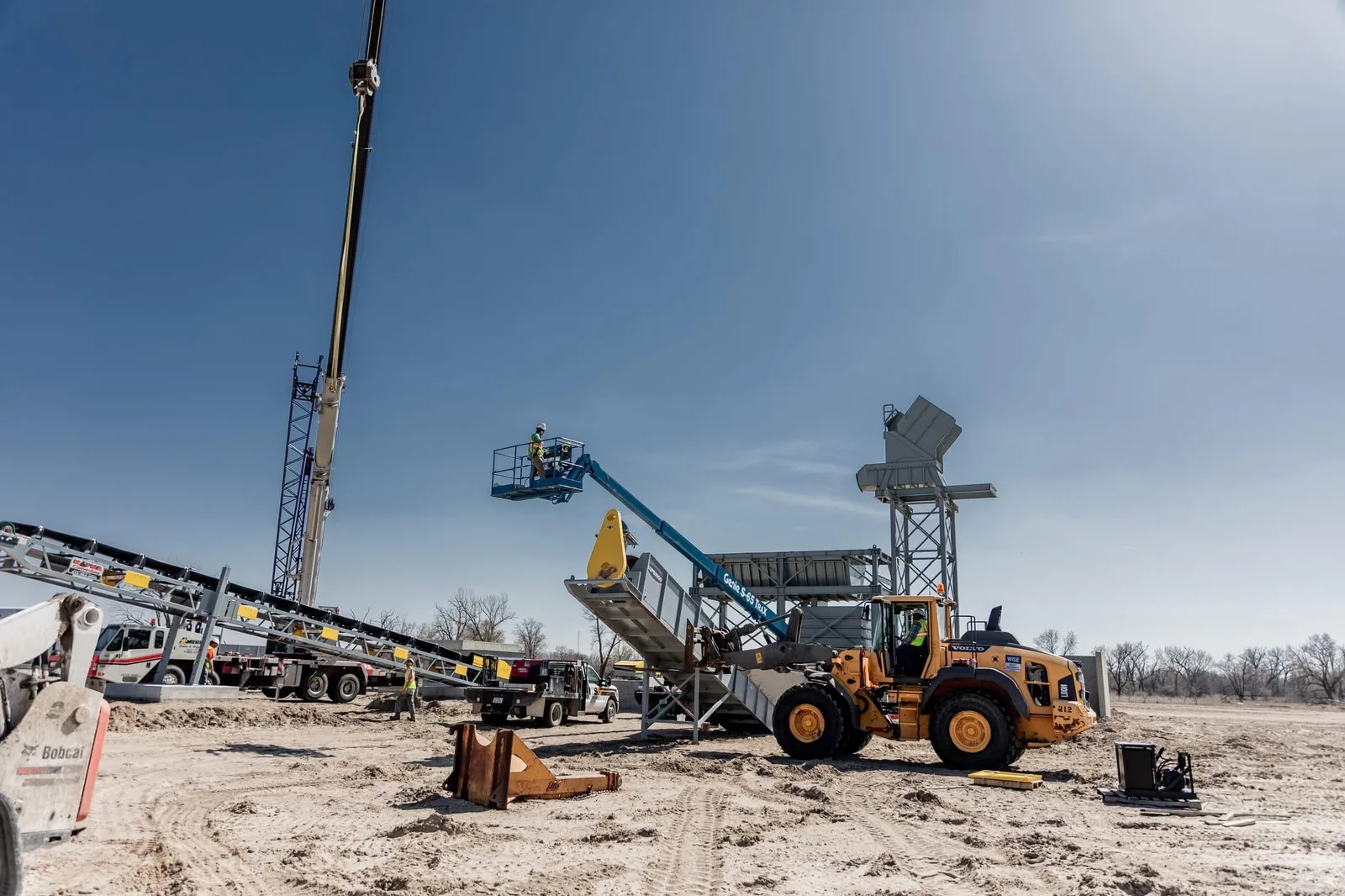 Wide jobsite shot with Bobcat skid steer, Volvo wheel loader, conveyor, and crew